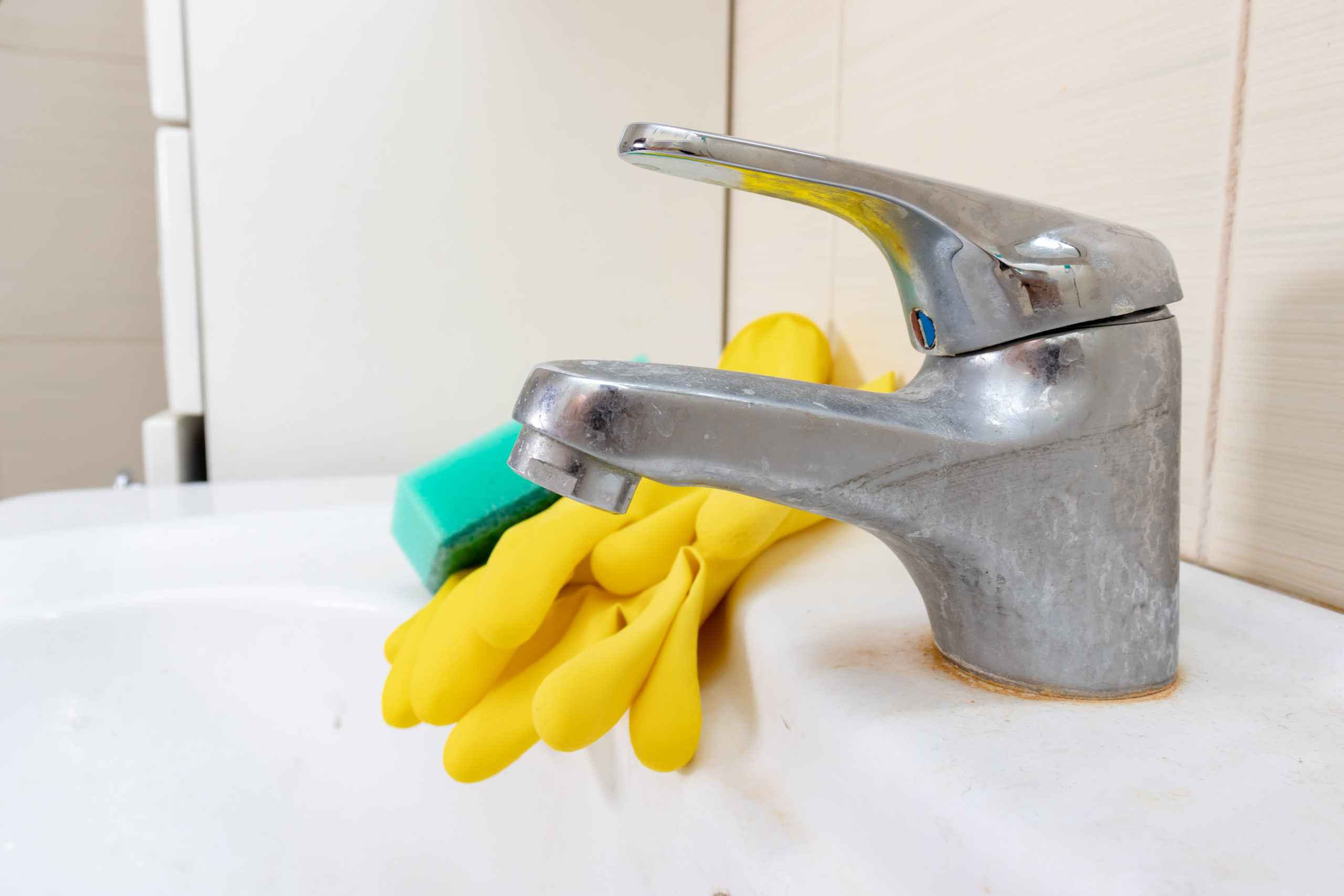 Verkalktes Waschbecken im Bad mit Gummihandschuhen und Schwamm Foto eines schmutzigen Bad-Waschbeckens mit Kalkablagerungen, worauf gelbe Gummihandschuhe und ein grüner Schwamm liegen.
