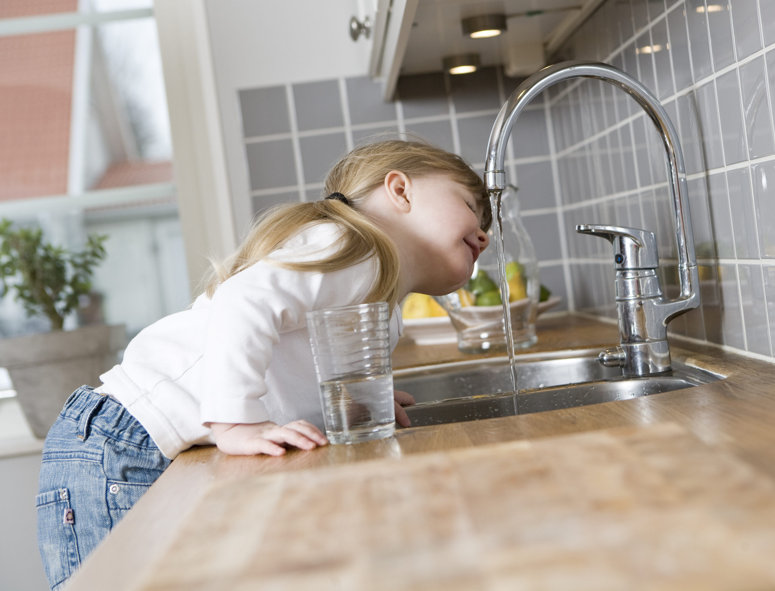 Reines Leitungswasser Kleines Mädchen trinkt Wasser direkt aus dem Wasserhahn in der Küche. Daneben steht ein halbvolles Wasserglas.