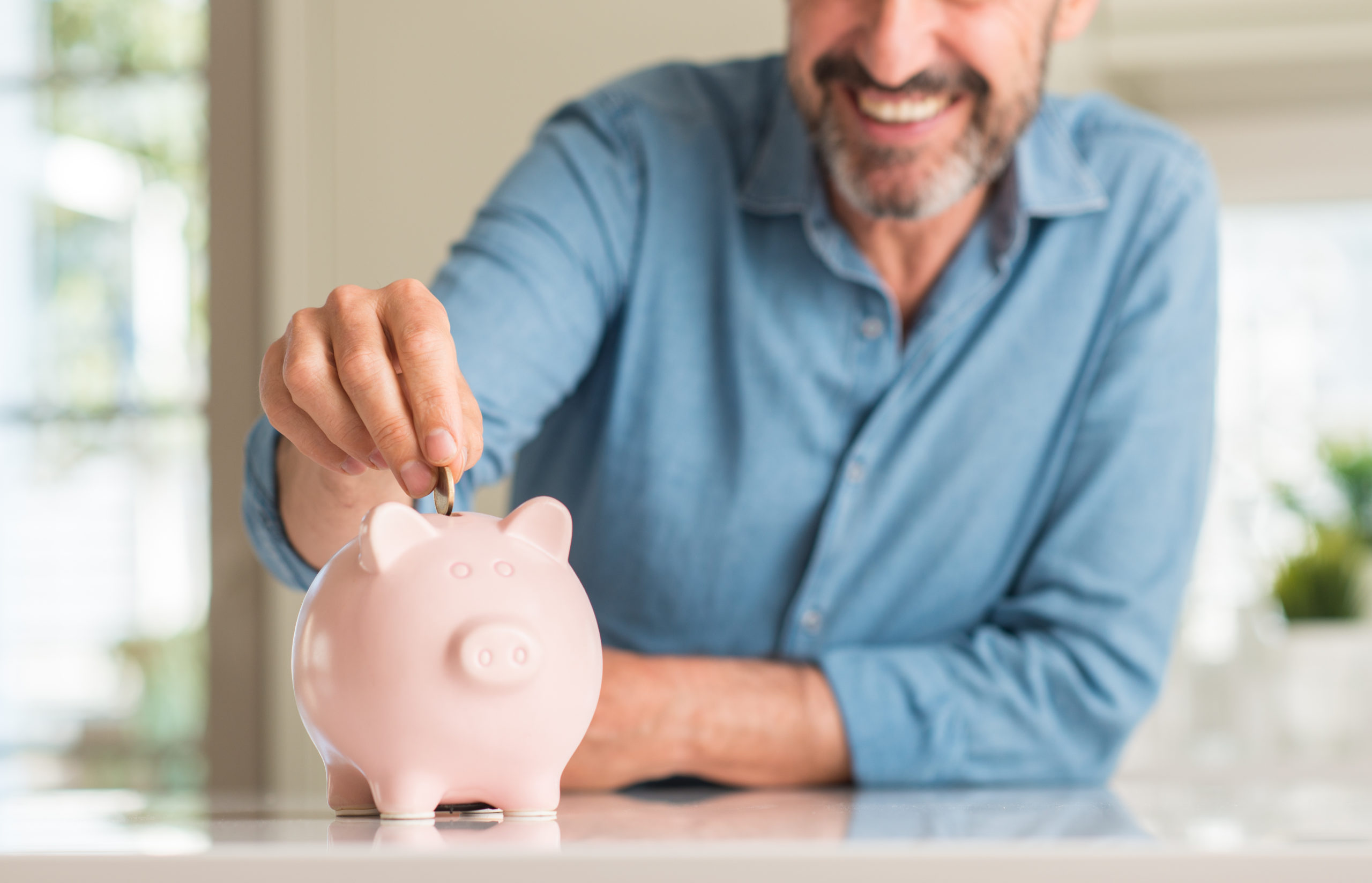 Mann im blauen Hemd und mittleren Alter spart Geld in einem rosa Sparschwein. Er lächelt glücklich und zufrieden.