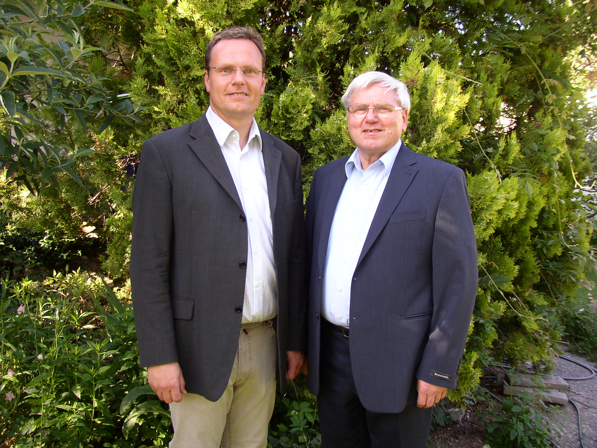 Hermann und Thomas Löhnert, die Gründer der Löhnert GmbH, stehen im Anzug vor einem grünen Busch